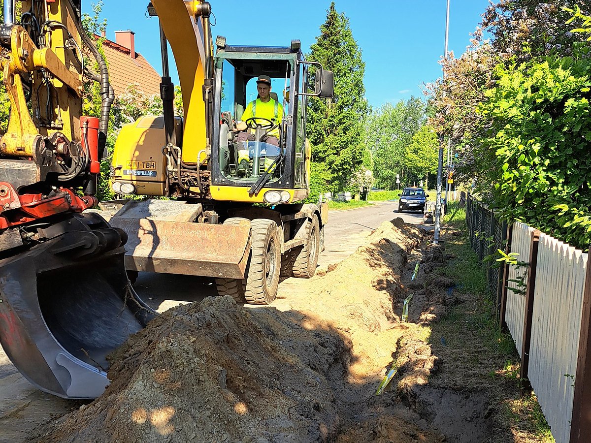 Pyöräalustainen kaivinkone kaivaa kaivantoa kadun reunaan, kuljettaja ohjaamossa